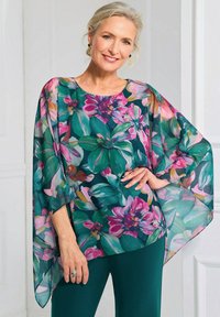 Blouse met bloemen in doorschijnende stof, met een groene achtergrond en grote roze en paarse bloemen. Losse, vloeiende snit met brede mouwen.
