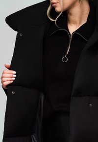 Blondýnka v černém svršku na zip a rozepnuté černé péřové bundě, ukazuje pečlivě upravené bílé nehty, jak drží bundu.