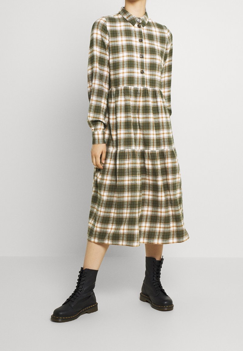 Robe à carreaux vert et blanc avec un col, manches longues et boutonnage sur le devant. Jupe évasée avec design à volants. Bottes noires à lacets.