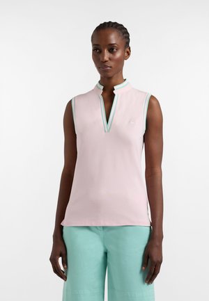 Vrouw met ingevlochten haar, gekleed in een mouwloos, lichtroze top met muntgroene rand en brede muntgroene broek, staand tegen een witte achtergrond.