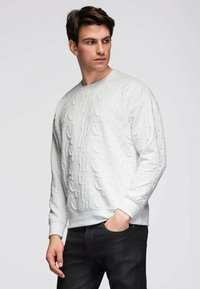 Jeune homme portant un sweat-shirt blanc à motifs embossés et un jean noir, regardant vers sa gauche avec une main dans la poche.