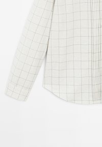Chemise à manches longues en tissu beige clair avec un motif en grille. Elle présente un ourlet arrondi et de simples détails aux poignets.