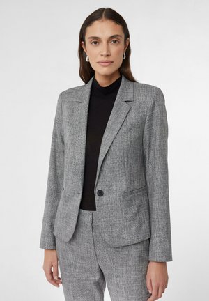 Blazer grigio con trama, colletto a revers, chiusura a un bottone e due tasche laterali, indossato sopra un dolcevita nero.