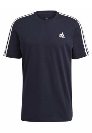 Mann trägt ein dunkelblaues Adidas-T-Shirt mit weißen Streifen auf den Schultern und graue Adidas-Hosen, steht vor einem schlichten Hintergrund.