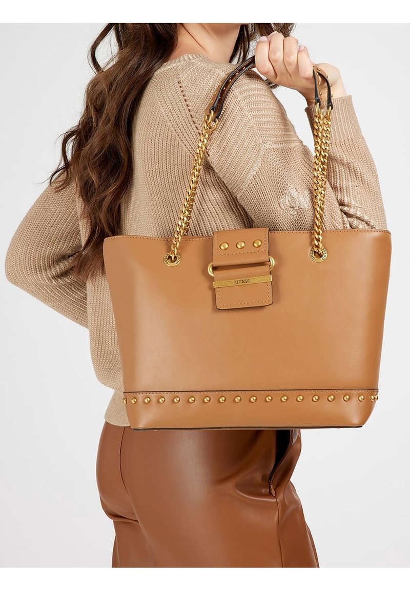 Donna che tiene una grande borsa a tracolla in pelle beige con manici in catena dorata e rifiniture borchiate, indossando un maglione beige e pantaloni marroni.