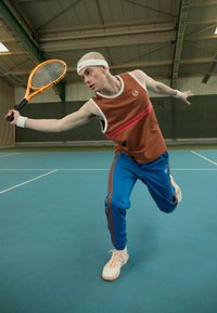 Sportlane, kes on riietatud pruunisse varrukateta särki koos punase triibuga, sinistesse jooksuhästi, millel on oranžid triibud, ja valgetesse kingadesse, hoides käes oranzi tennise reketit.