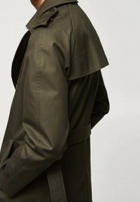 Olivgrüner Trenchcoat mit strukturiertem Design, der über einen Klappkragen, Schulterklappen und eine gebundene Taille verfügt, aus einem glatten Material gefertigt.