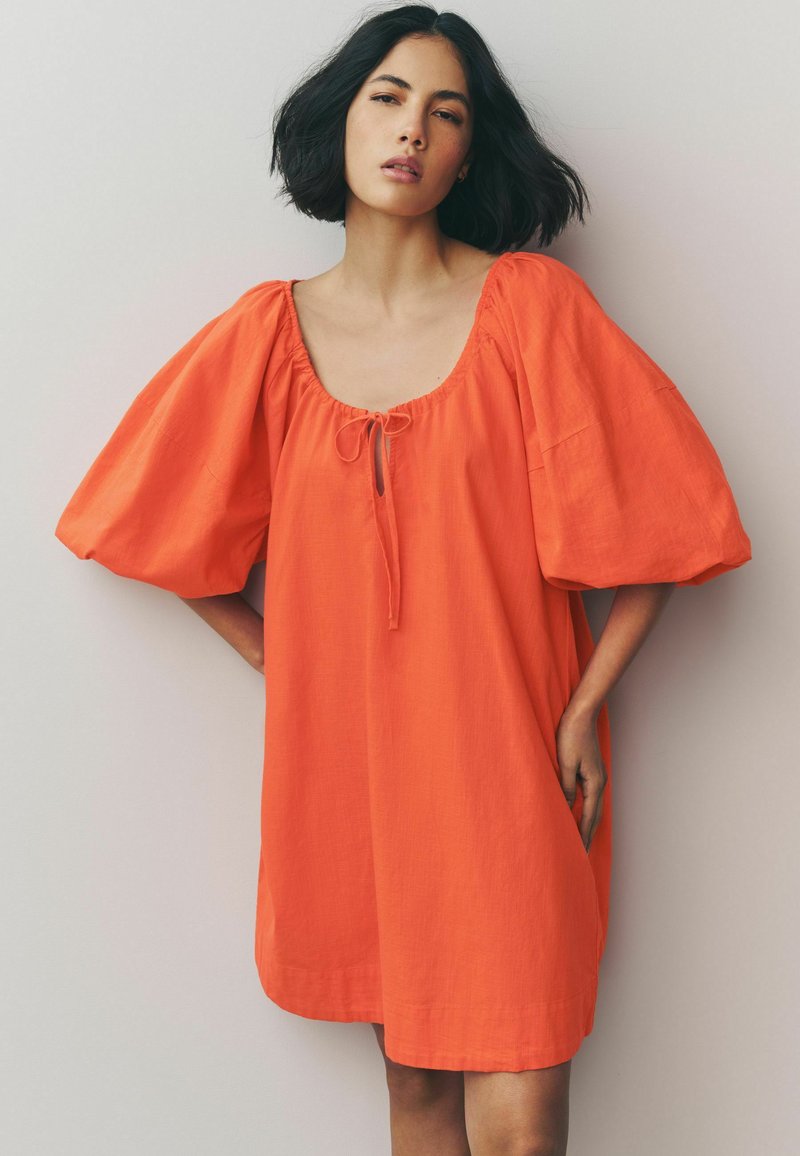 Korte oranje linnen jurk met een losse pasvorm, brede mouwen en een vierkante halslijn met een strikdetail in het midden. Glad textuur.