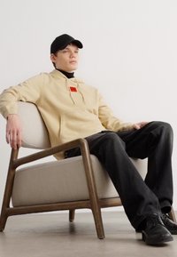 Sudadera beige con un logo rojo, cuello alto negro, jeans negros y zapatos negros. Sentado en una silla gris claro con patas de madera.