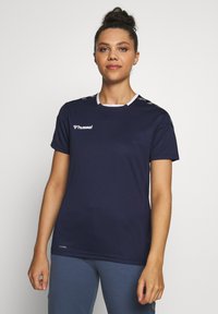 Giovane donna con i capelli raccolti in uno chignon che indossa una maglietta sportiva Hummel blu navy a maniche corte e pantaloni azzurri, in piedi davanti a uno sfondo bianco uniforme.