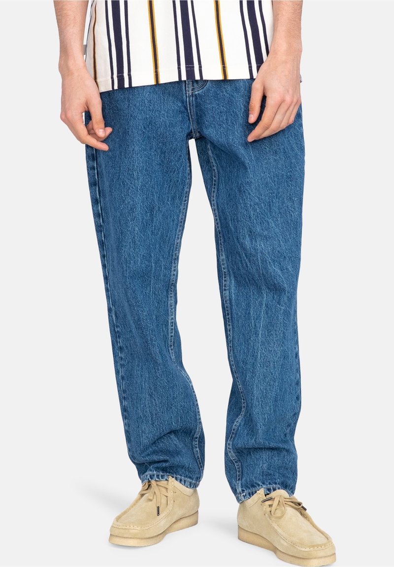 Jean en denim bleu clair à coupe décontractée, avec un léger effet délavé et des détails de couture. Porté avec une chemise blanche à rayures et des chaussures beiges.