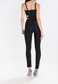 Kvinna i en svart ärmlös jumpsuit med utskärningar och dragkedja på ryggen, matchad med silverfärgade högklackade skor, stående mot vit bakgrund.