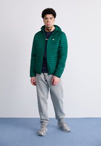 Zelená puffer bunda so zipsom a kapucňou, nosená cez námornícku modrú blúzku, kombinovaná so šedými teplákmi a striebornými športovými topánkami.