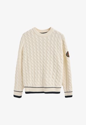 Maglione a trecce color crema con collo girocollo a costine, orlo a strisce blu e una toppa sul manica sinistra con un design decorativo.