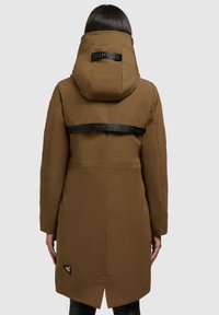 Manteau isolé marron avec un col haut, présentant une sangle de logo noire dans le dos et un patch de marque près de l'ourlet.