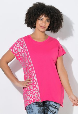 Kvinde med krøllet hår iført en knaldpink kortærmet top med sølv leopardprint og lynlåsdetaile, står med den ene hånd på hoften.