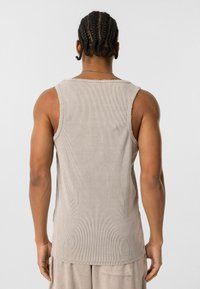 Geribbelde, mouwloze taupe tanktop met een ruwe zoom, met een relaxte pasvorm en subtiele textuur. Model wordt van achteren getoond.