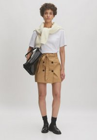 Jupe beige à double boutonnage avec des boutons noirs et un nœud sur le devant, associée à un t-shirt blanc et une écharpe crème texturée, portant un sac noir.