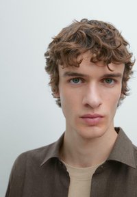 Portræt af en person med bølget brunt hår, iført en cremefarvet skjorte under en brun skjorte med knapper. Fokus på ansigtstræk og udtryk.