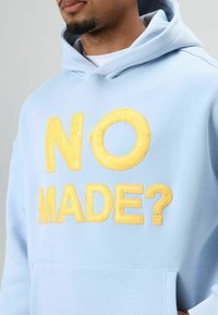 Hoodie bleu clair en tissu doux avec un grand texte brodé jaune « NO MADE ? » sur le devant et une poche kangourou.