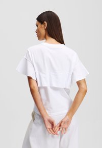Tricou alb din bumbac cu un design stratificat în spate, mâneci scurte și evazate și guler clasic rotund. Textură netedă, fără modele.