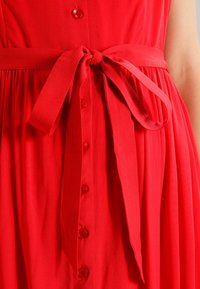 Robe rouge avec des boutons sur le devant et une ceinture en tissu nouée à la taille, montrée du milieu du torse jusqu'aux hanches.