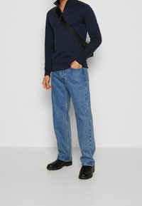 Uomo che indossa un maglione blu navy con zip a quarto girocollo, jeans blu a gamba dritta, stivali neri e borsa a tracolla nera, in piedi con una mano in tasca.