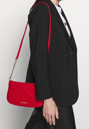 Red quilted crossbody bag with a chain strap, featuring a smooth texture and a rectangular shape. Gold hardware logo accent visible.