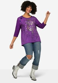 Lila Langarmshirt mit Rundhalsausschnitt und metallic Grafiken, kombiniert mit ausgewaschenen blauen Jeans und silbernen Stiefeletten.