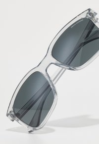 Ochelari de soare cu rame din plastic transparent și lentile gri închis. Formă pătrată și linii curate, având logo-ul pe partea interioară a ramei.
