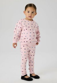 Małe dziecko stojące, ubrane w jasnoróżowy sweter i legginsy z fioletowymi i niebieskimi wzorami serc oraz czarne buty wsuwane, na jednolitym jasnym tle.