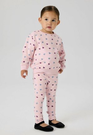 Tout-petit debout, portant un pull rose pâle et des leggings avec des motifs de cœurs violets et bleus, ainsi que des chaussures plates noires, sur un fond clair uni.