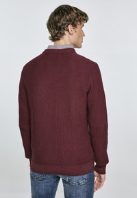 Burgunderfarbener Pullover mit strukturiertem Strickmuster und gerippten Bündchen. Kombiniert mit einem karierten Hemd darunter, das einen klassischen Kragen hat.