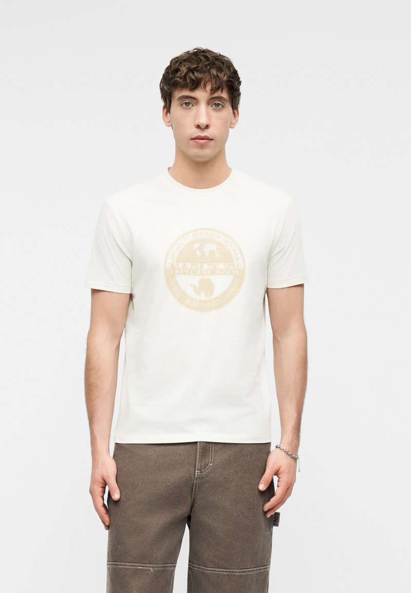 Νέος άνδρας με καστανά σγουρά μαλλιά, φορώντας λευκό μπλουζάκι με γραφικά και γκρι παντελόνι, στέκεται μπροστά σε απλό φωτεινό φόντο.