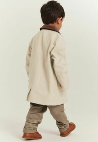 Beige trenchcoat med brun krage, bärs över olivgröna byxor. Bruna skor med mjuk textur. Enkel design, inga synliga mönster.
