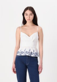 Witte peplum top met een V-hals en dunne bandjes, versierd met blauwe bloemenborduursels aan de onderkant. Gecombineerd met blauwe denim jeans.