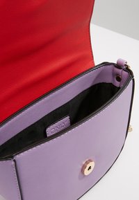 Fialová kožená taška s červenou podšívkou, černým lemováním, magnetickým zavíráním a malou značkovou štítkem uvnitř, se jemnou texturou.