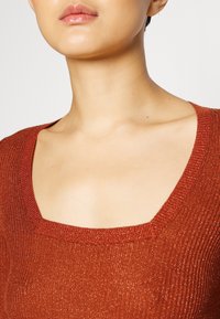 Gros plan du haut du torse et du cou portant un pull côtelé de couleur rouille avec un décolleté carré et des fils métalliques discrets.