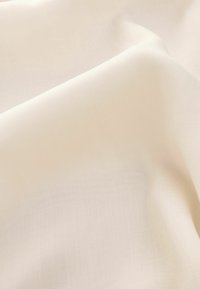 Světle krémová tkanina s hladkou, jemnou texturou, zobrazená ve skladech, které vykazují jemný lesk a jemný vzor tkaní. Žádné viditelné vzory ani kovové prvky.