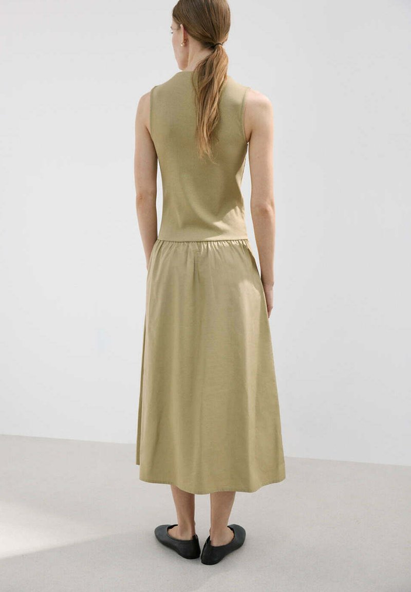 Femme aux longs cheveux attachés en queue de cheval, portant un haut beige sans manches, une jupe midi assortie et des chaussures plates noires, debout sur un sol clair contre un mur uni.