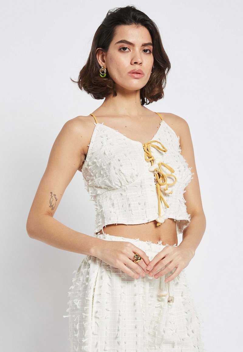 Top bianco corto con texture sfilacciata, lacci gialli regolabili e accenti decorativi. Abbinato a una gonna sfilacciata coordinata.