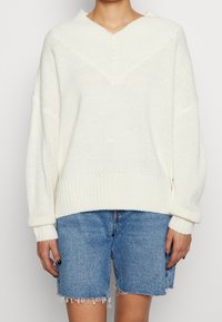 Weißer Strickpullover mit V-Ausschnitt und überschnittenen Schultern. Gerippte Bündchen und Saum, mit lockerer Passform. Kombiniert mit Jeans-Shorts.