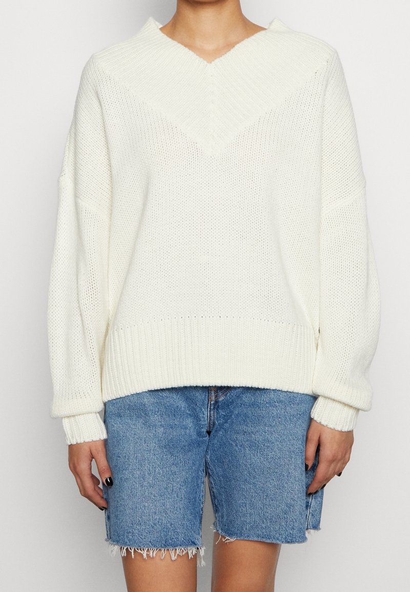 Weißer Strickpullover mit V-Ausschnitt und überschnittenen Schultern. Gerippte Bündchen und Saum, mit lockerer Passform. Kombiniert mit Jeans-Shorts.