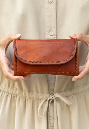 Braune Lederbörse mit einer geschwungenen Klappe, sichtbaren Nähten und rechteckiger Form. In Händen gegen einen neutralen Stoffhintergrund gehalten.