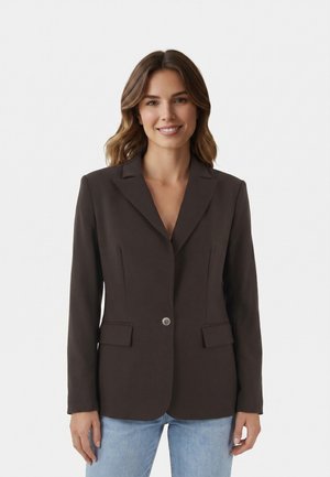 Vrouw met middellang bruin golvend haar draagt een donkere single-breasted blazer en lichtblauwe jeans, staand en glimlachend tegen een egale achtergrond.