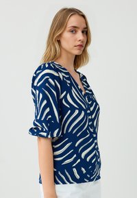 Camicia blu navy con motivi a onda bianchi, tessuto testurizzato, scollo a V e maniche corte a sbuffo. Presenta chiusura frontale con bottoni e vestibilità aderente.