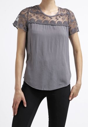 Blouse grise avec empiècement et manches en dentelle florale transparente, portée avec un pantalon noir, mains posées sur les hanches.