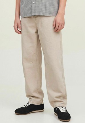 Nedre del af en person iført beige løstsiddende bukser og sorte sneakers med hvide snørebånd, stående mod en ensfarvet lys baggrund.