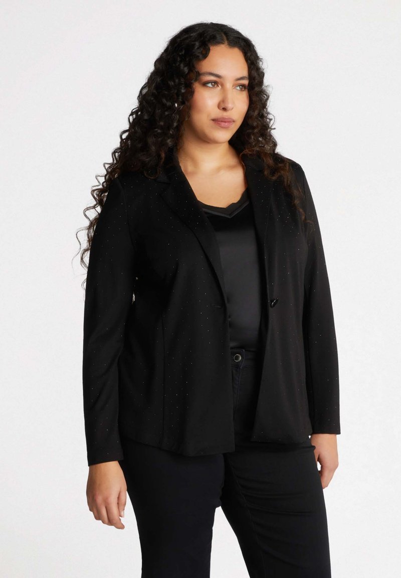 Svart kavaj med slagkrage, subtildottad mönster, lättviktsmaterial, enda knappstängning och en avslappnad passform. Bärs över en svart topp.