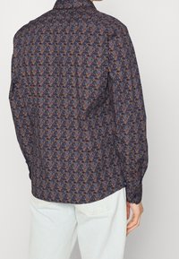Homme portant une chemise à manches longues avec un motif floral sombre, assortie à un jean bleu clair, vu de dos sur un fond uni.
